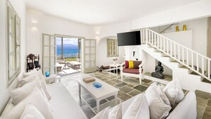A spacious living room with a white sofa, a coffee table, and a staircase leading to an upper floor. A large flat-screen TV is mounted on the wall, and a statue is placed on the staircase landing.