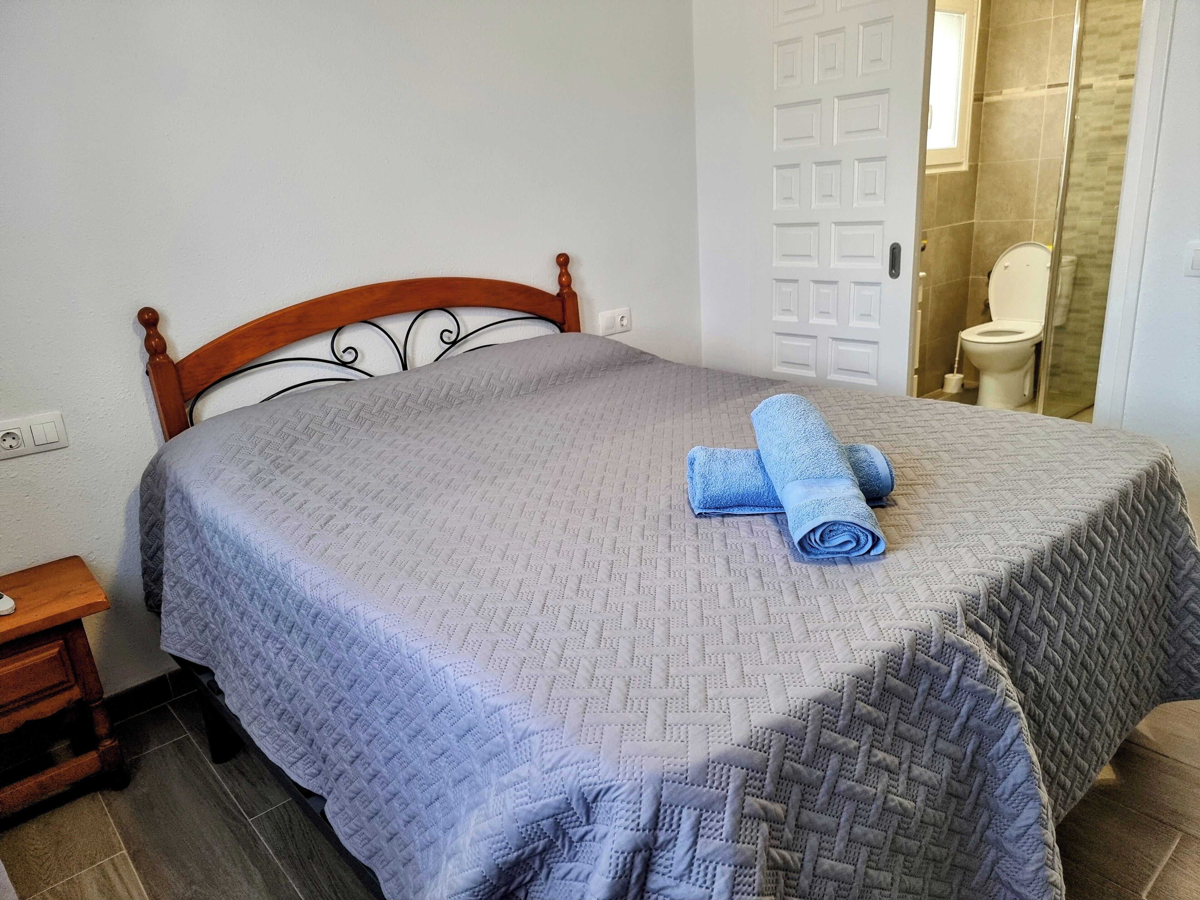 A neatly made bed with a grey quilt and rolled blue towels, a wooden headboard, a nightstand with a lamp, and a bathroom with a glass shower door.
