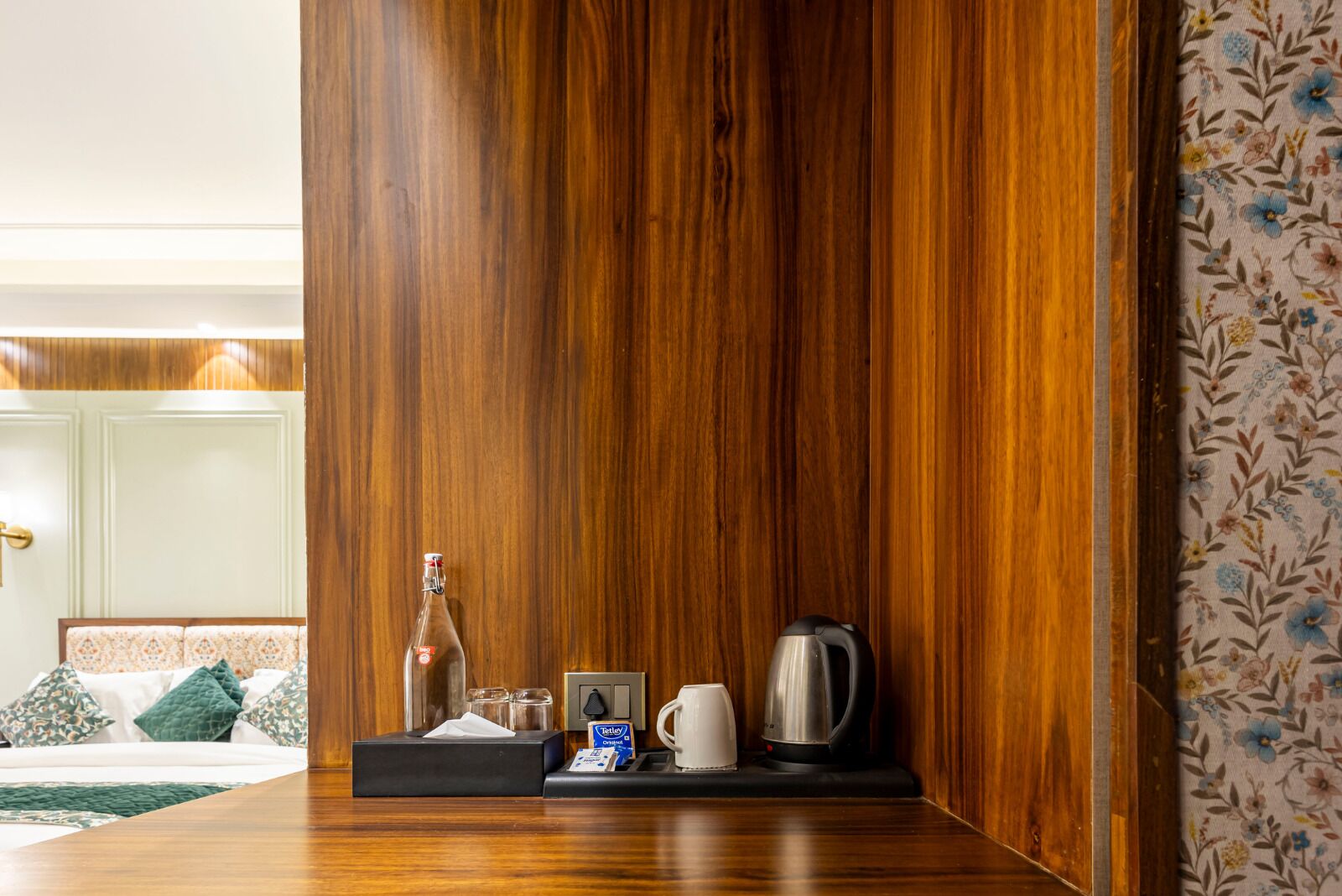 Habitación de hotel con pared de paneles de madera, mesita de noche con una botella, vasos, caja de pañuelos, hervidor y bandeja con una taza.