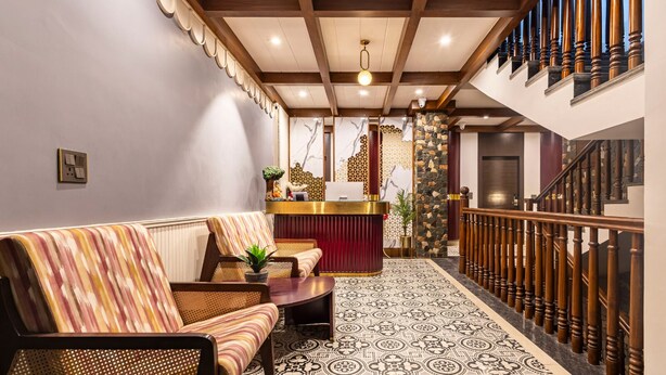 A reception area with a reception desk, a sofa, a patterned rug, and a staircase with wooden railings. - The Nathuli Manor (Mussoorie)