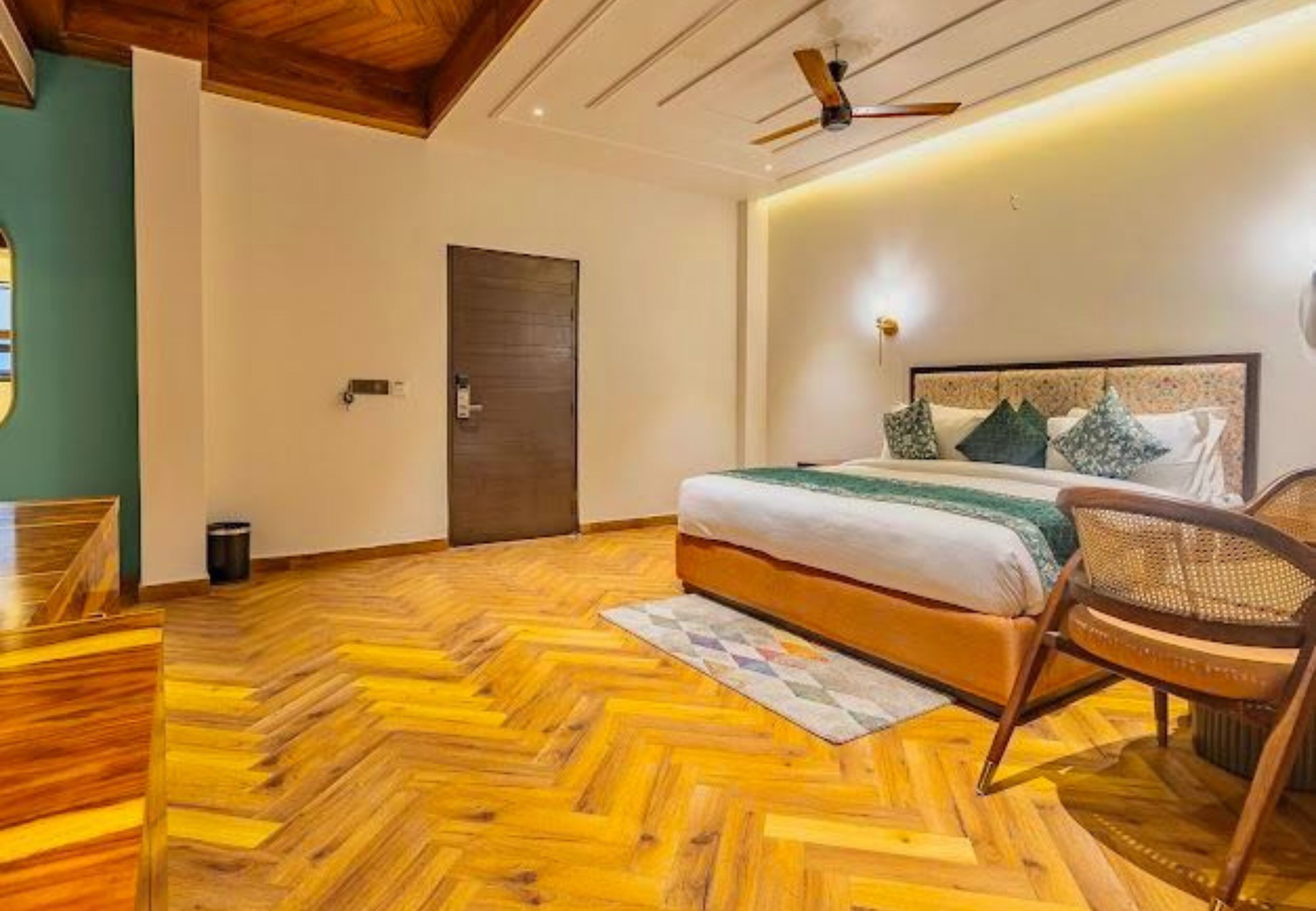A bedroom with a wooden herringbone floor, a bed with a green blanket, a wooden chair, and a ceiling fan.