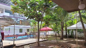 Área de piscina com um edifício branco, guarda-sol vermelho e árvores.