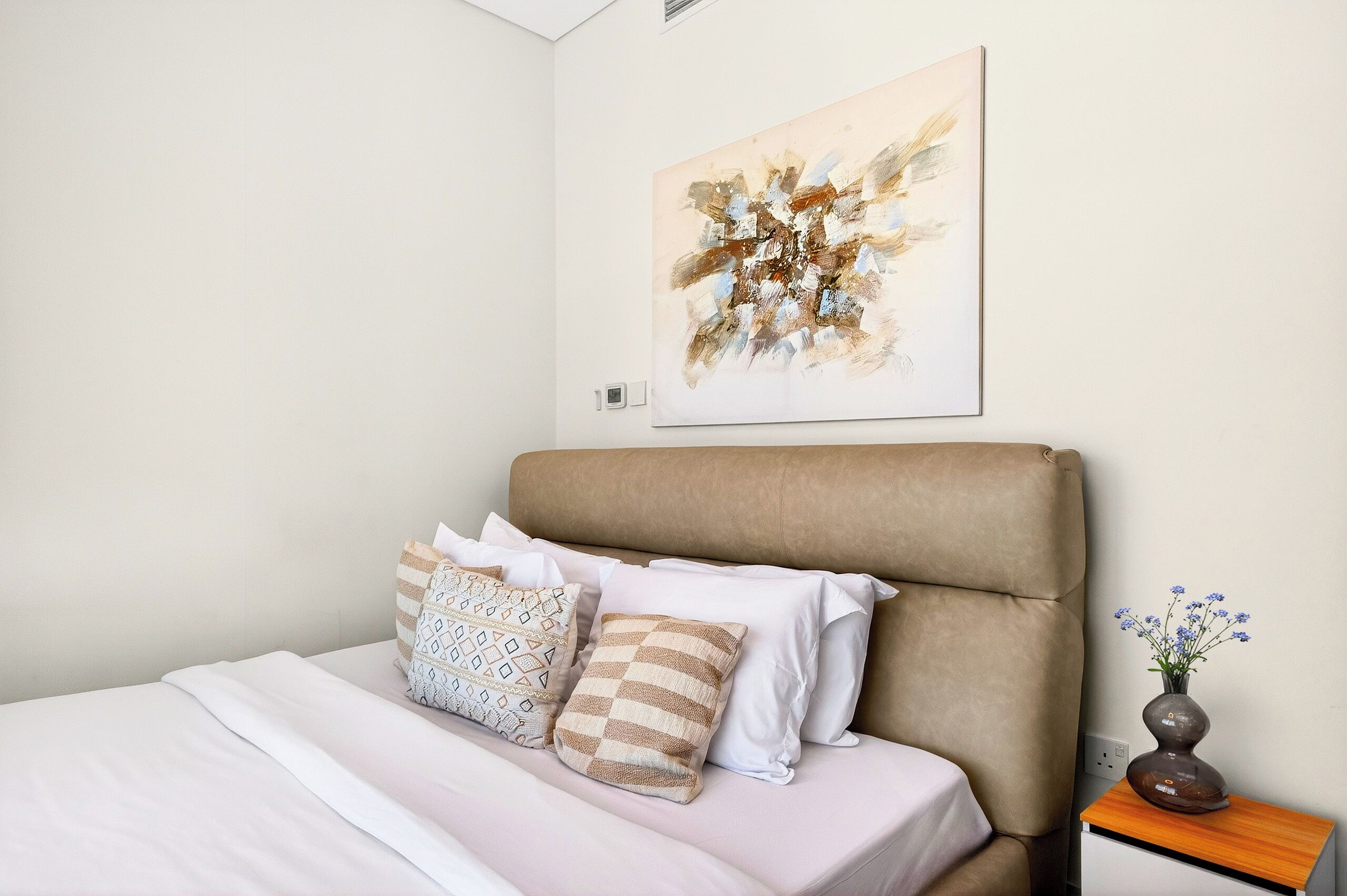 Una cama bien tendida con almohadas estampadas, un cabecero color beige, un cuadro abstracto colgado en la pared y una mesita de noche de madera con un florero.