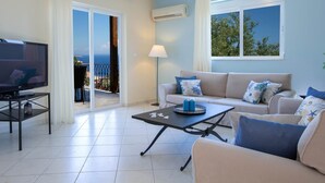 A living room with a TV, sofa, and coffee table, a balcony with a view, and an air conditioning unit. - Villa Eione-Thaleia - Lefkada Vacation (LEFKADA)