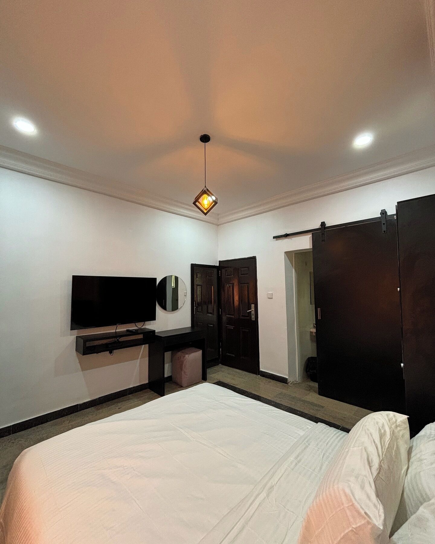 A hotel room with a large bed, a flat-screen TV mounted on the wall, a small stool, a wardrobe, and a door leading to another room.