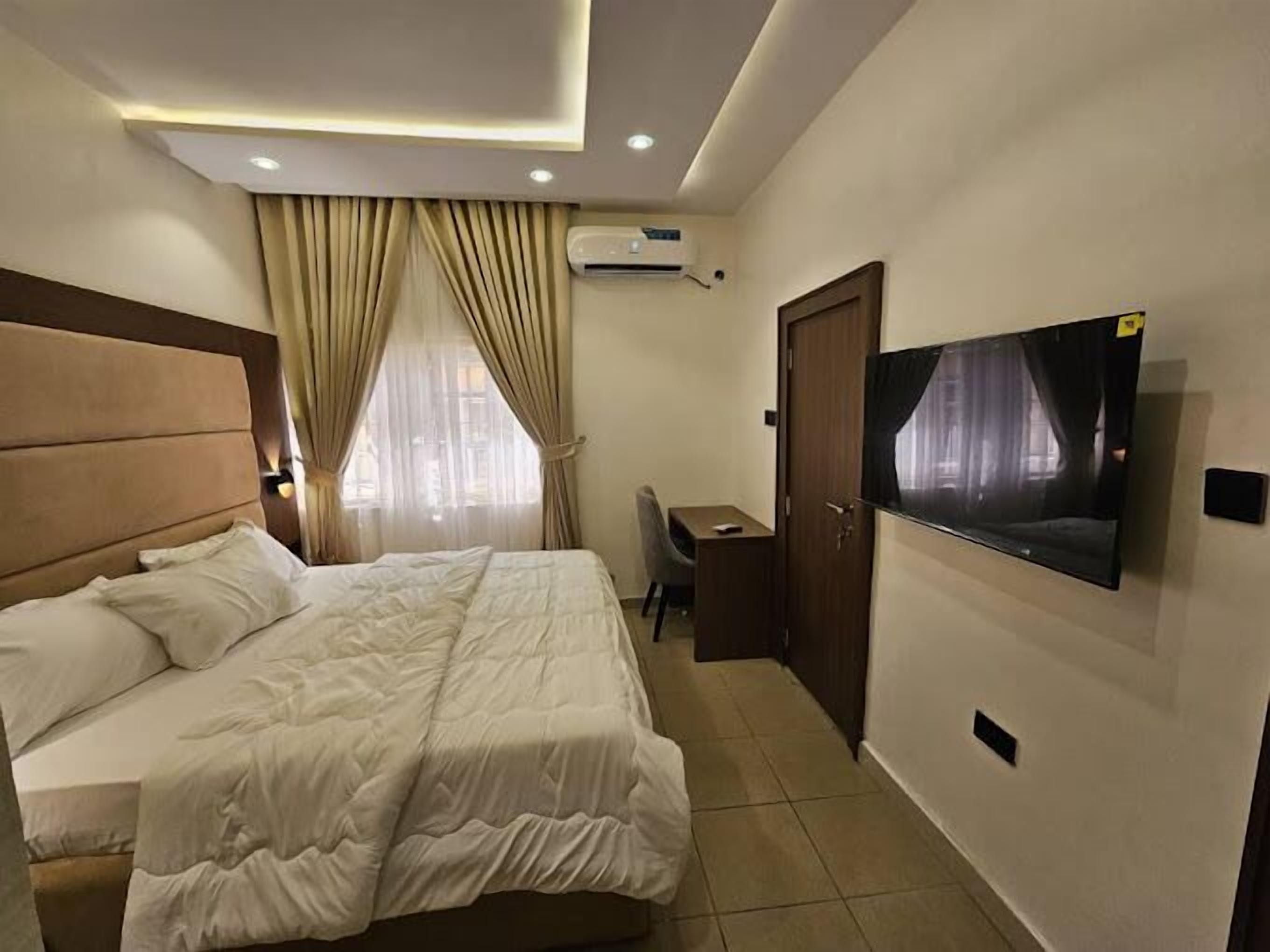 A hotel room with a bed, a desk, a chair, a TV mounted on the wall, and an air conditioning unit.
