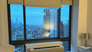 Quarto de hotel moderno com uma grande janela oferecendo vista para a cidade, uma cama, uma cadeira e uma mesa de cabeceira com abajur.