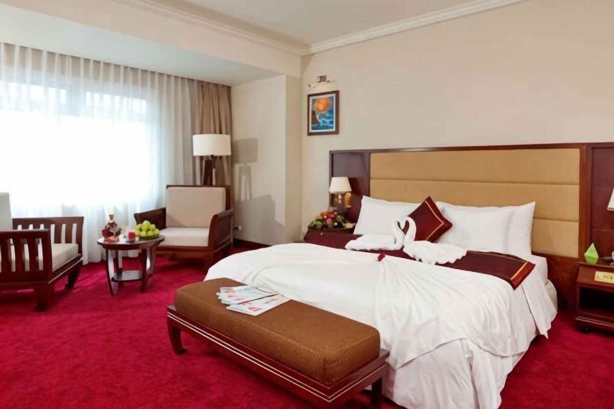 A hotel room with a large bed, a desk, a chair, a small table with fruit, and a window with curtains.