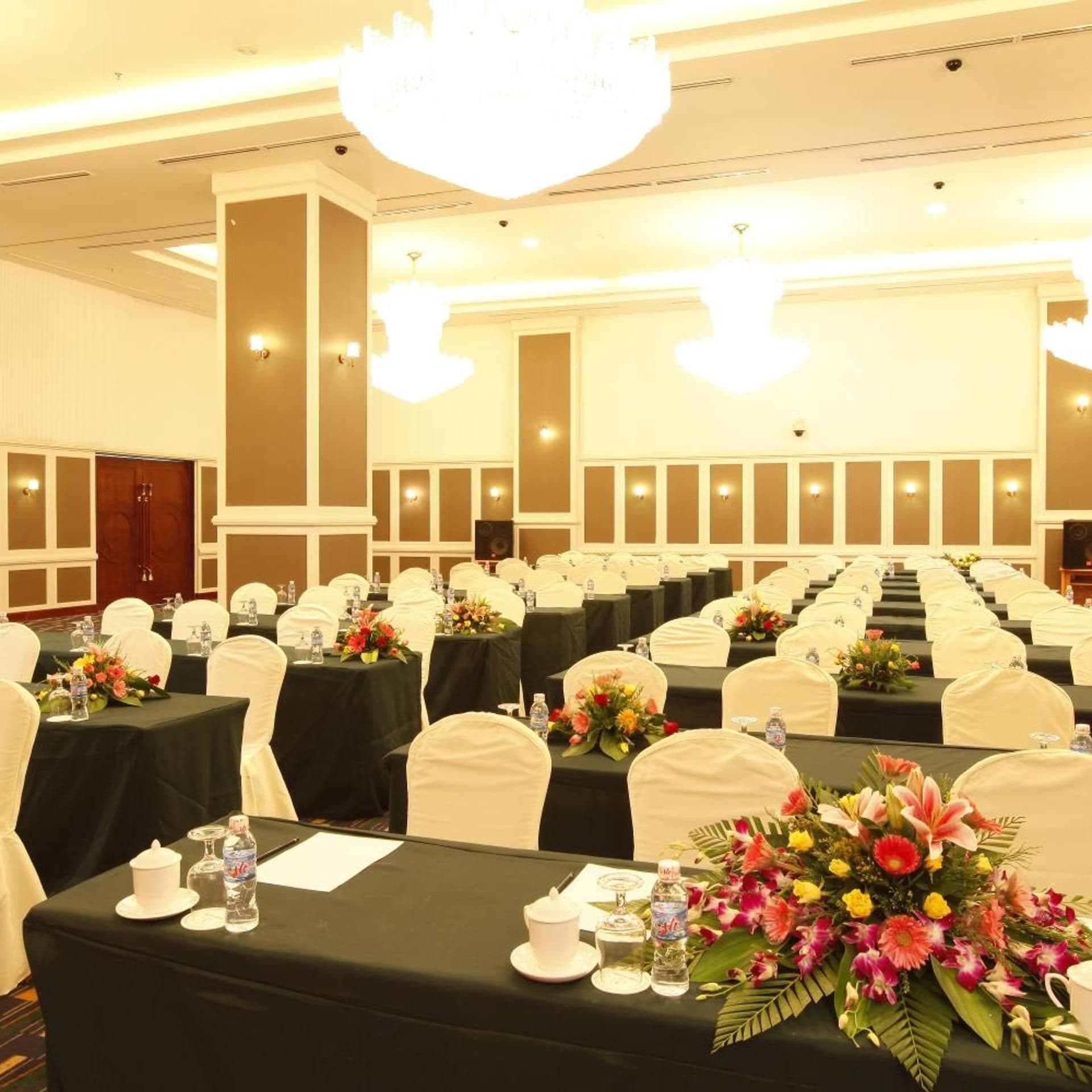 Un salón de banquetes con filas de mesas, cada una con una botella de agua, un vaso y un centro de mesa con flores.