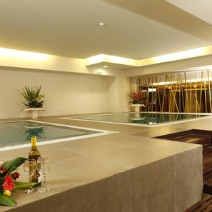 Un'area piscina interna moderna con pareti di vetro trasparente, una zona relax con una bottiglia di vino e bicchieri, e una scala che conduce a un altro livello.