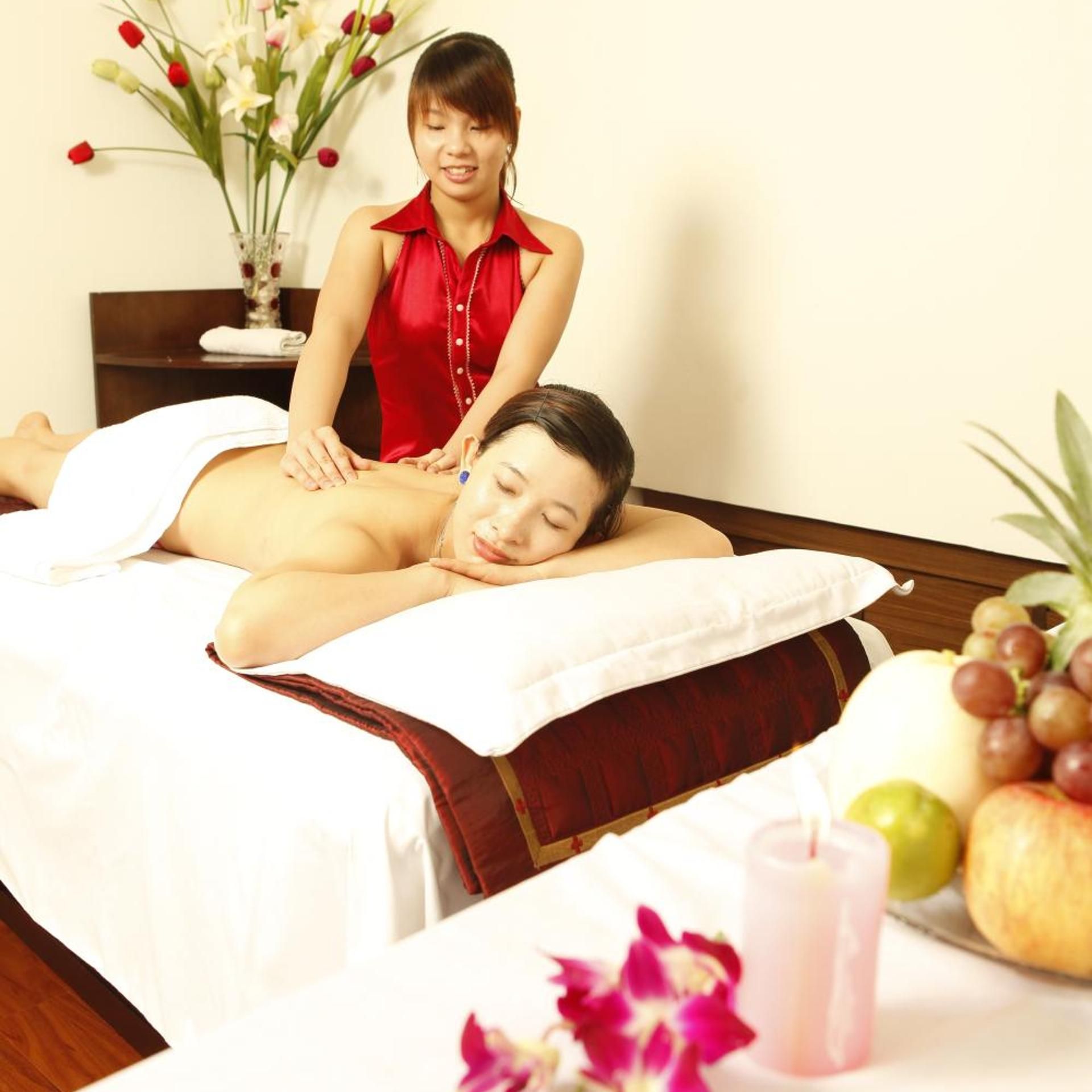A massage therapy session in a room with a bed, a massage table, a small table with fruits and a candle, and a vase with flowers.
