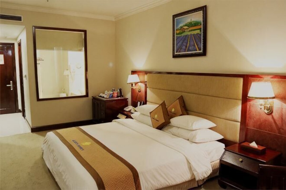 A hotel room with a large bed, two bedside tables, a nightstand, a mirror, and a painting on the wall.