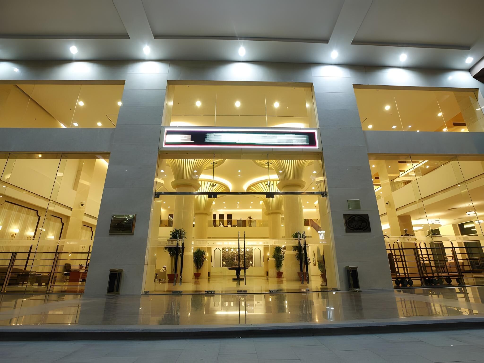 A modern hotel lobby with a central atrium, glass railings, and a digital signboard.