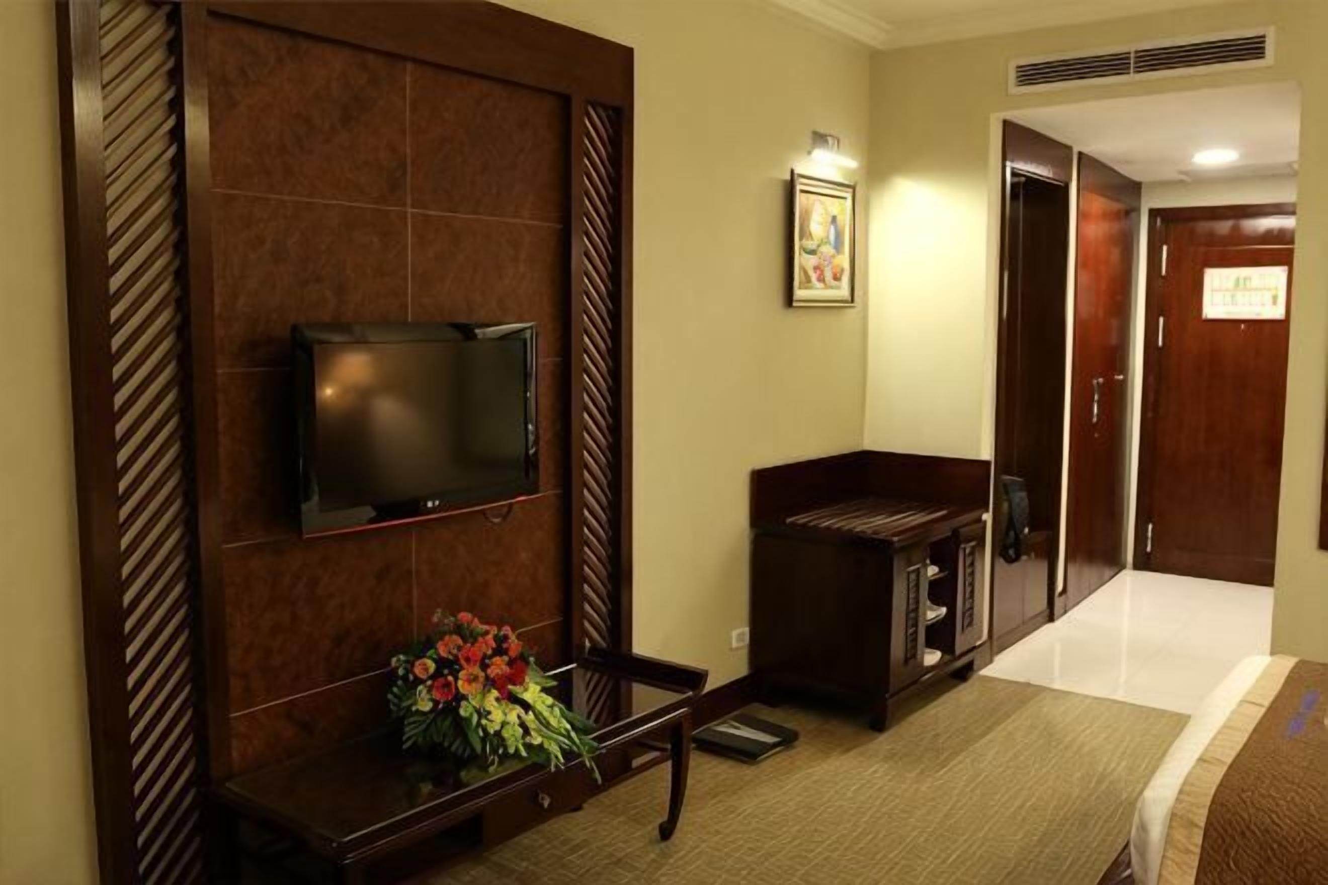 A hotel room with a flat-screen TV mounted on the wall, a wooden entertainment unit, a small table with a vase of flowers, and a door leading to another room.