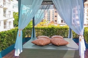 Un espacio de estar al aire libre cubierto con una cama y cojines, rodeado de follaje y edificios.
