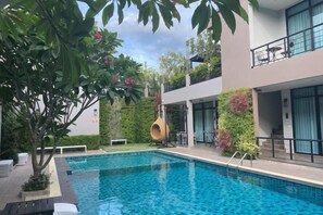 A swimming pool with a modern building in the background, a hanging chair, and a well-maintained garden. - Pearl Villa (Hua Hin)