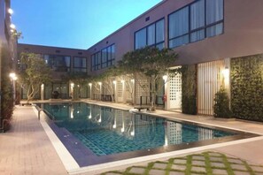 Una piscina con solárium de azulejos, rodeada por un edificio con grandes ventanales y techo plano.