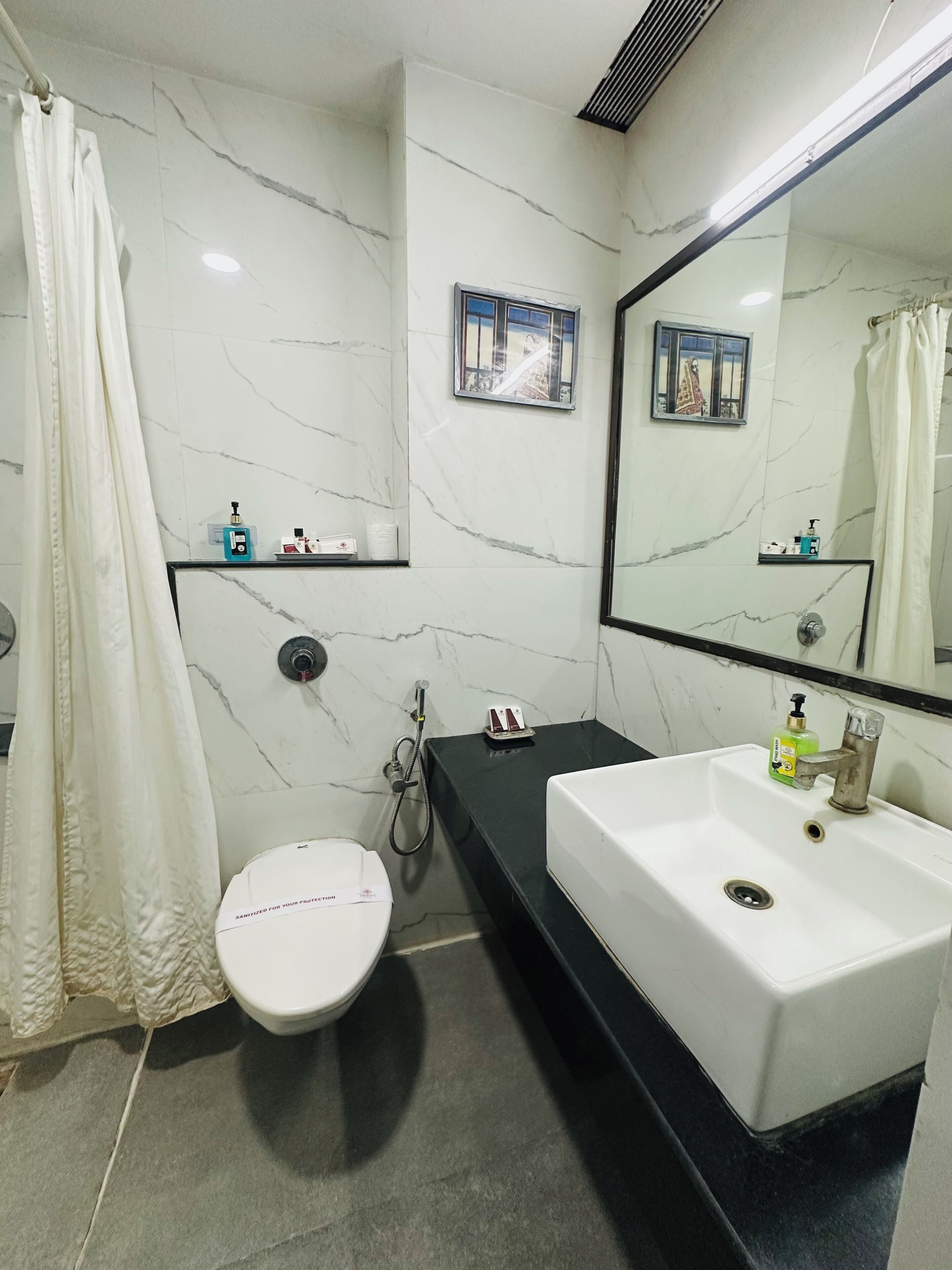 A bathroom with a white toilet, a black vanity with a white sink, a shower area, and a mirror with a picture frame.
