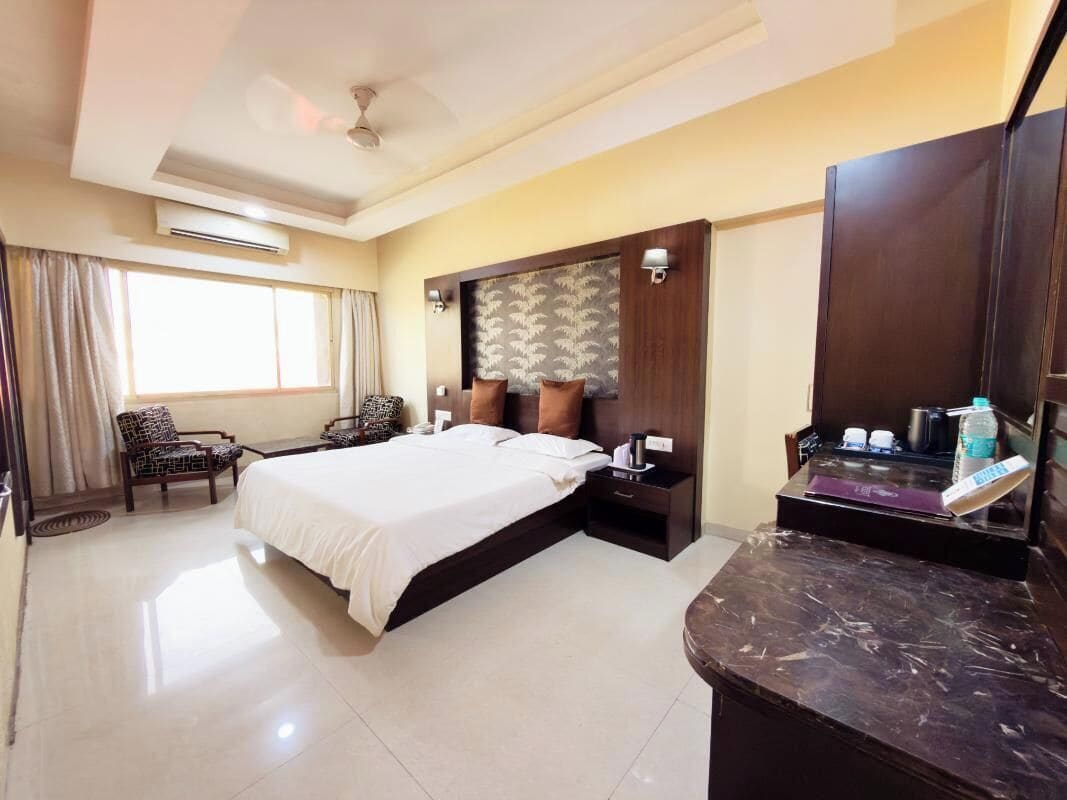 Quarto de hotel com cama, duas cadeiras, escrivaninha, penteadeira e uma janela grande.