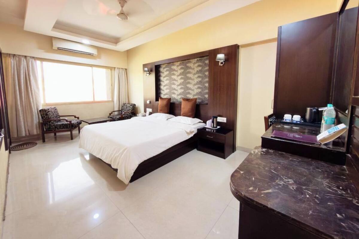 Une chambre d’hôtel avec un lit, deux chaises, un bureau, une coiffeuse et une grande fenêtre.