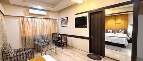 Habitación de hotel con cama, televisor, escritorio con sillas, sofá y un espejo grande.