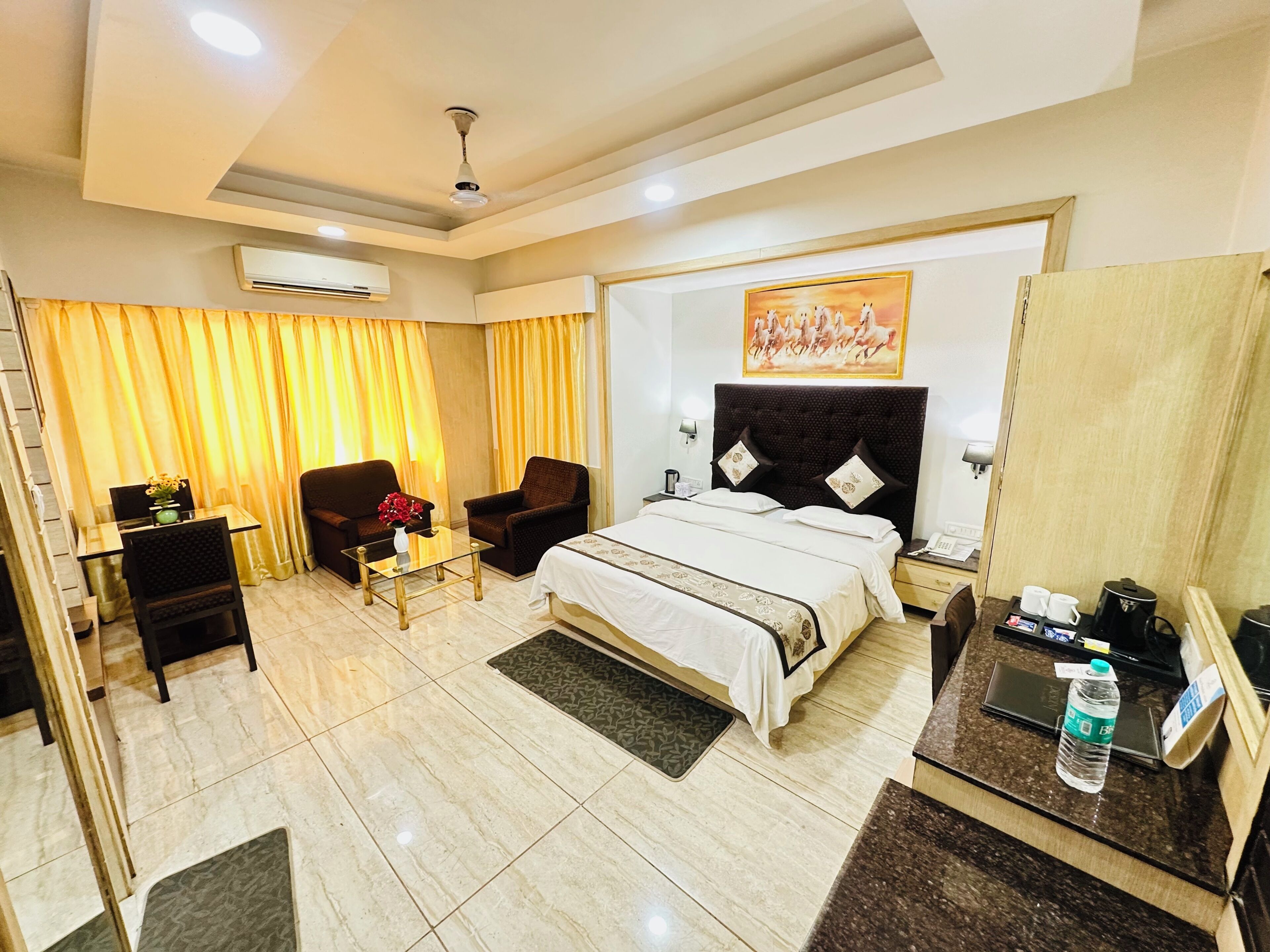 Quarto de hotel com cama, duas poltronas, uma mesinha de centro com um vaso, uma escrivaninha com telefone e um quadro na parede.