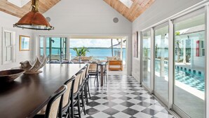 A spacious dining area with a long table, chairs, and a large window overlooking a pool and ocean view.