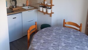 A compact kitchen with white cabinets, a wooden shelf with jars, a sink, and a dining area with a patterned tablecloth. - Joli studio face à la mer avec accès direct (Saint-Cyprien)
