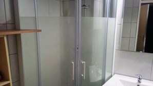 A corner shower enclosure with a glass door, a white sink, and a mirror above it. - Joli studio face à la mer avec accès direct (Saint-Cyprien)