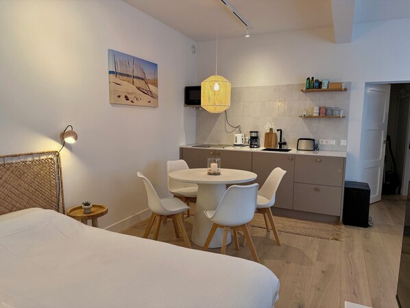 A modern hotel room with a bed, a dining table with chairs, a kitchen area with a coffee maker, and a painting on the wall.