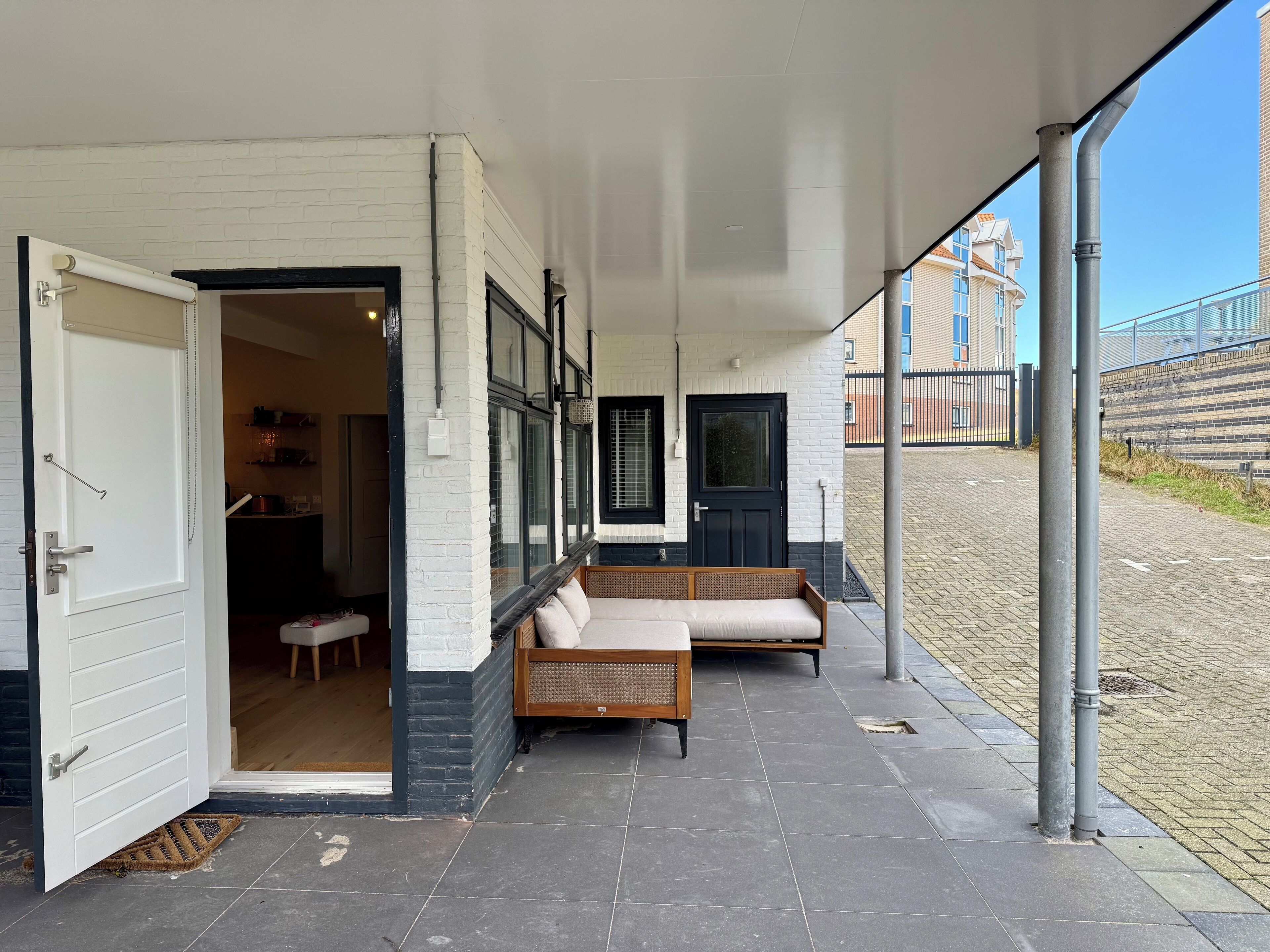 A covered outdoor seating area with a sofa and a bench, a door leading to an interior space, and a clear view of the street and buildings in the background.