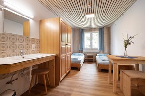 A room with a wooden floor, a wooden bed, a wooden desk, a wooden chair, a wooden wardrobe, a wooden table, a wooden bench, a wooden stool, a wooden cabinet, a wooden sink, a mirror, a window with curtains, a radiator, and a plant.