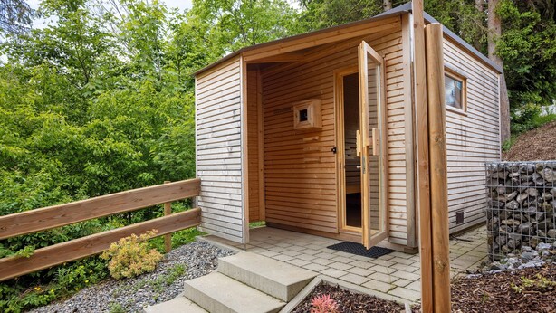 Eine Holzhütte mit geöffneter Tür, umgeben von einem Garten mit Stufen, die zu ihr hinaufführen.