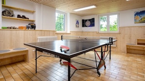 Ein Pingpongtisch mit Netz, Holzregale mit Schuhen und Taschen sowie eine Dartscheibe an der Wand.