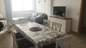 Zona de menjador amb taula preparada per a dues persones, un sofà, una televisió i una finestra amb persianes.