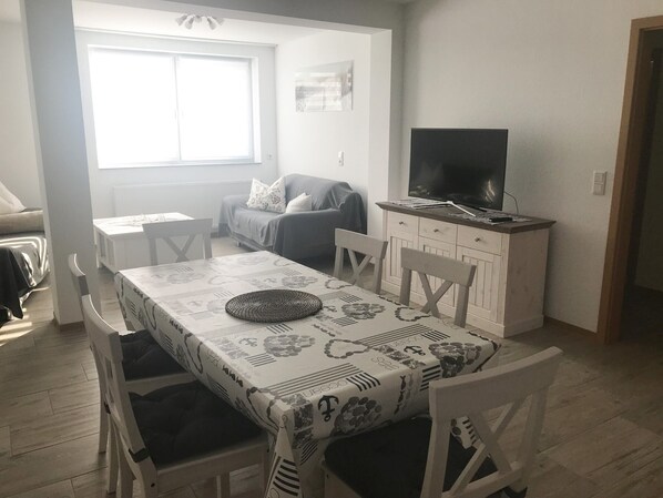 A dining area with a table set for two, a sofa, a television, and a window with blinds.