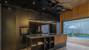 A modern kitchen with built-in appliances, a wooden accent wall, and a view of a swimming pool. - Villa Sunset Berawa (Canggu)