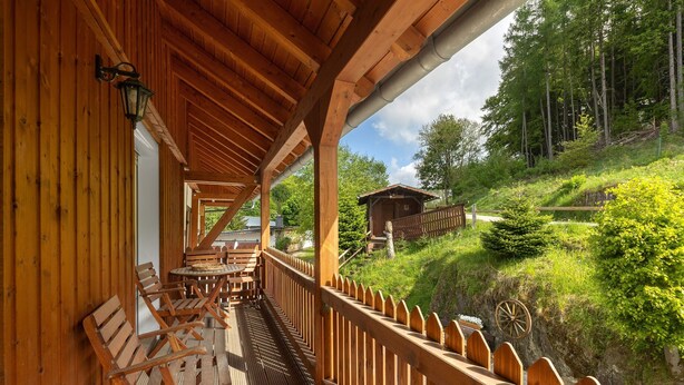 Une terrasse en bois avec des sièges d’extérieur, une vue sur une colline boisée et un bâtiment rustique en arrière-plan.