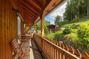 A wooden deck with outdoor seating, a view of a forested hillside, and a rustic building in the background.