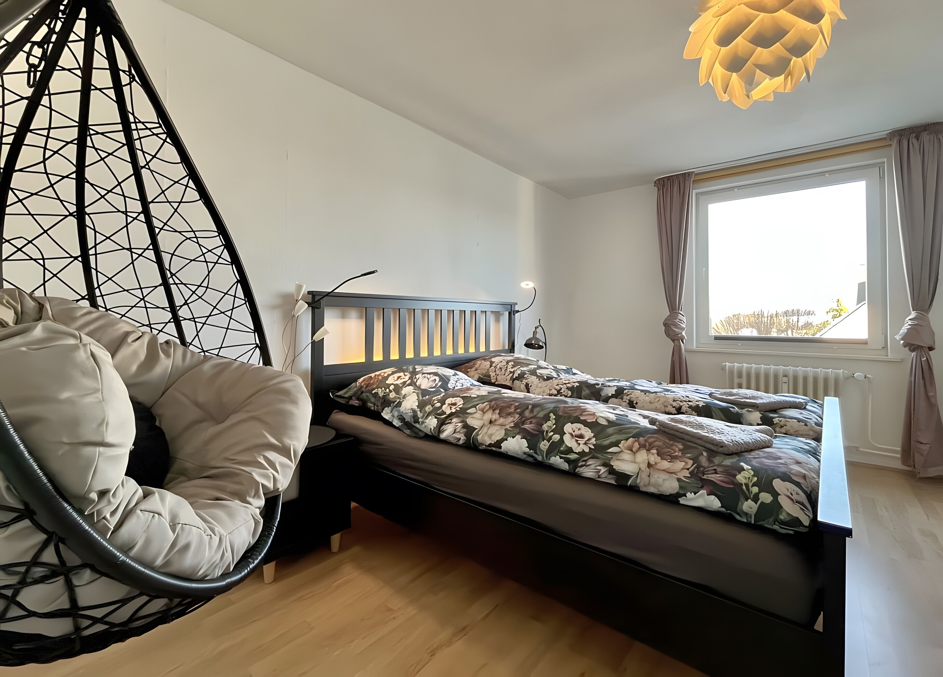 A bed with floral bedding, a hanging chair, a bedside lamp, and a window with curtains.