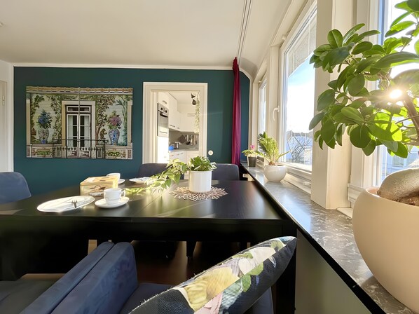 A dining area with a table set for two, a potted plant, and a view of the outside.
