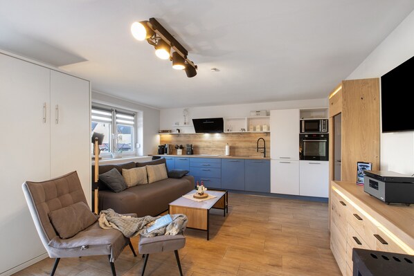 A modern living room with a sofa, armchair, and coffee table, a kitchen with blue cabinets and a microwave, and a TV mounted on the wall.