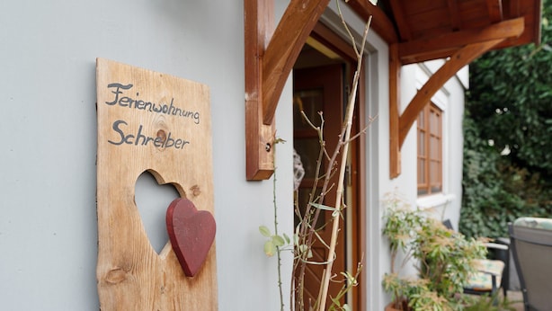 Een houten bord met een hartvormig gat en tekst, een houten constructie en een bloempot met plant.