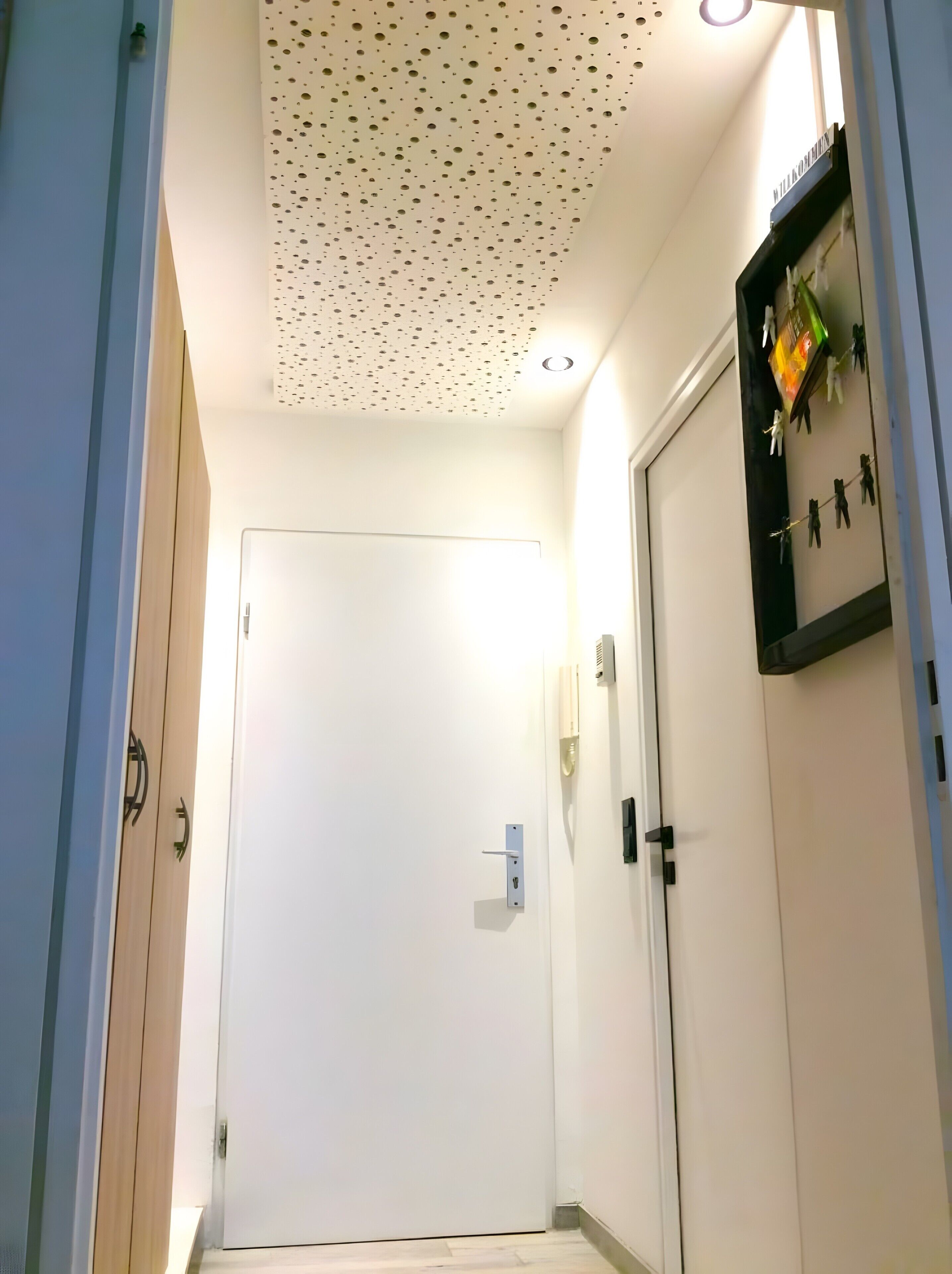 Un pasillo con una puerta blanca, techo con diseño perforado y una lámpara fijada a la pared.