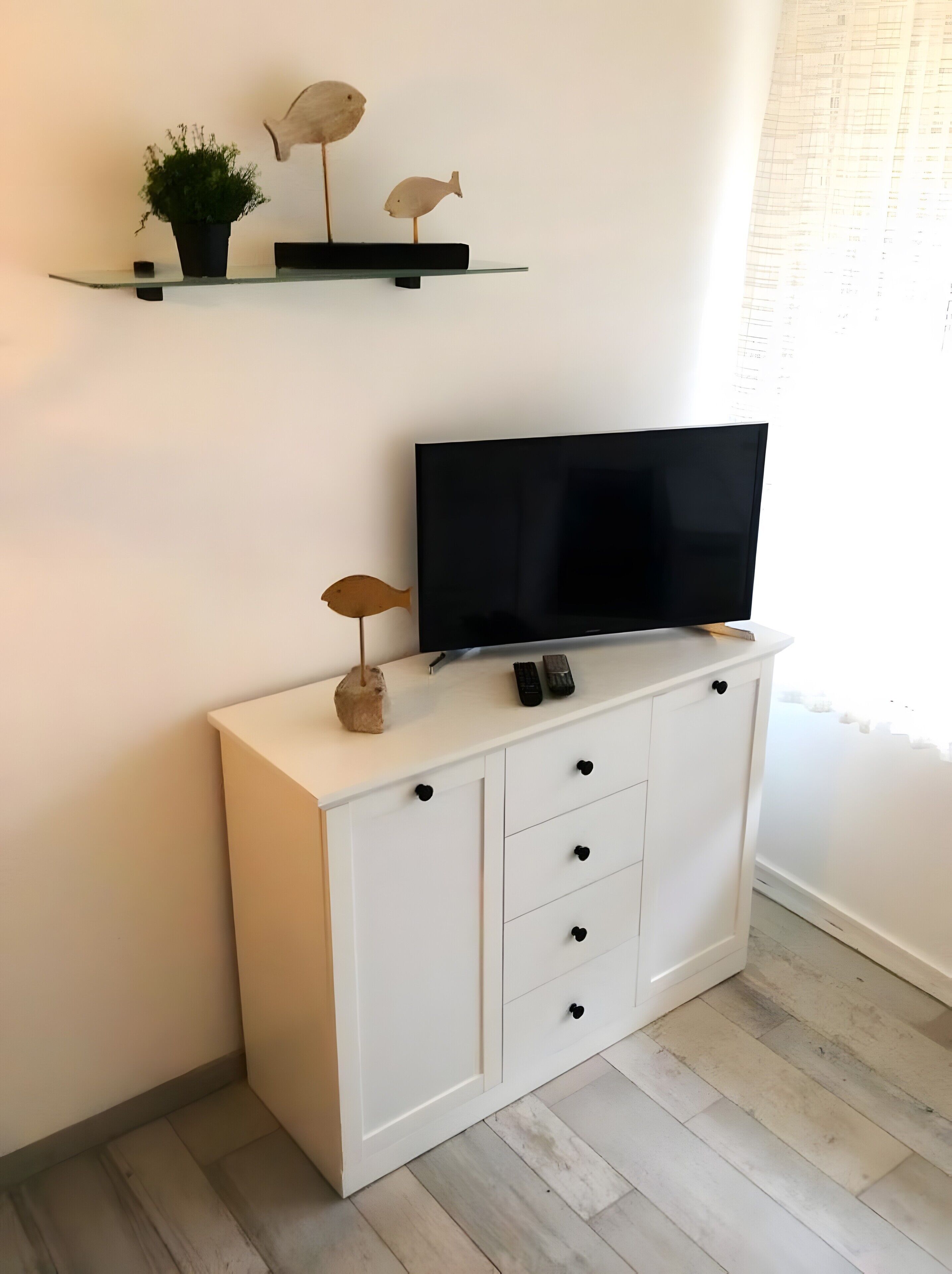 A white cabinet with a flat-screen TV, a small potted plant, and two wooden fish decorations on a shelf above.