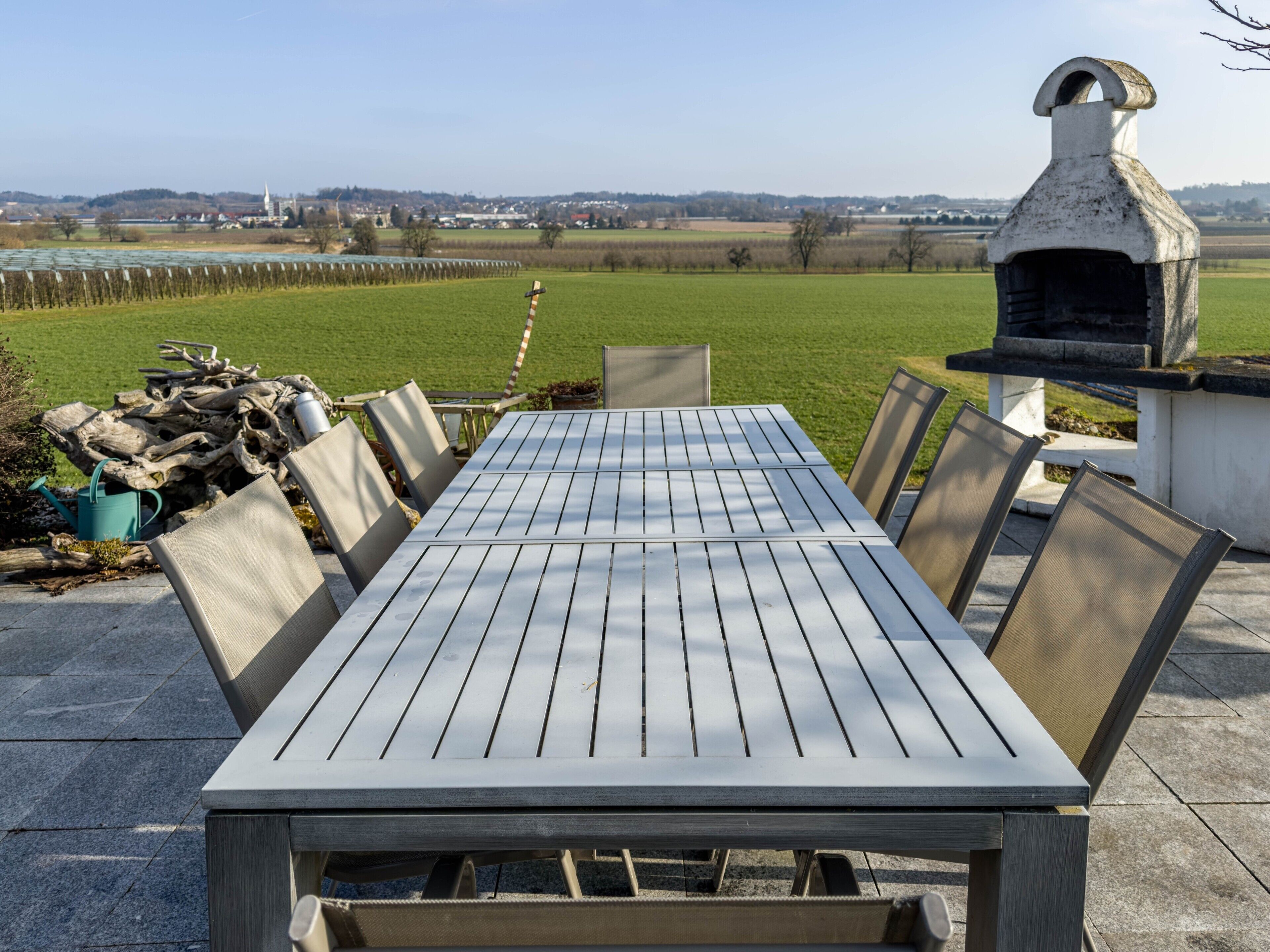Eine Terrasse im Freien mit Tisch und Stühlen, einem rustikalen Grill und Blick auf ein grünes Feld sowie entfernte Gebäude.