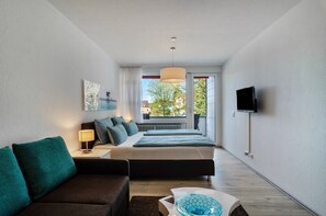 Habitación de hotel moderna con cama, sofá, televisión y ventana con vistas a edificios y árboles.