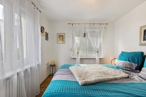 A bedroom with a bed, a window with sheer curtains, a bedside table with a candle, and a wall-mounted picture.