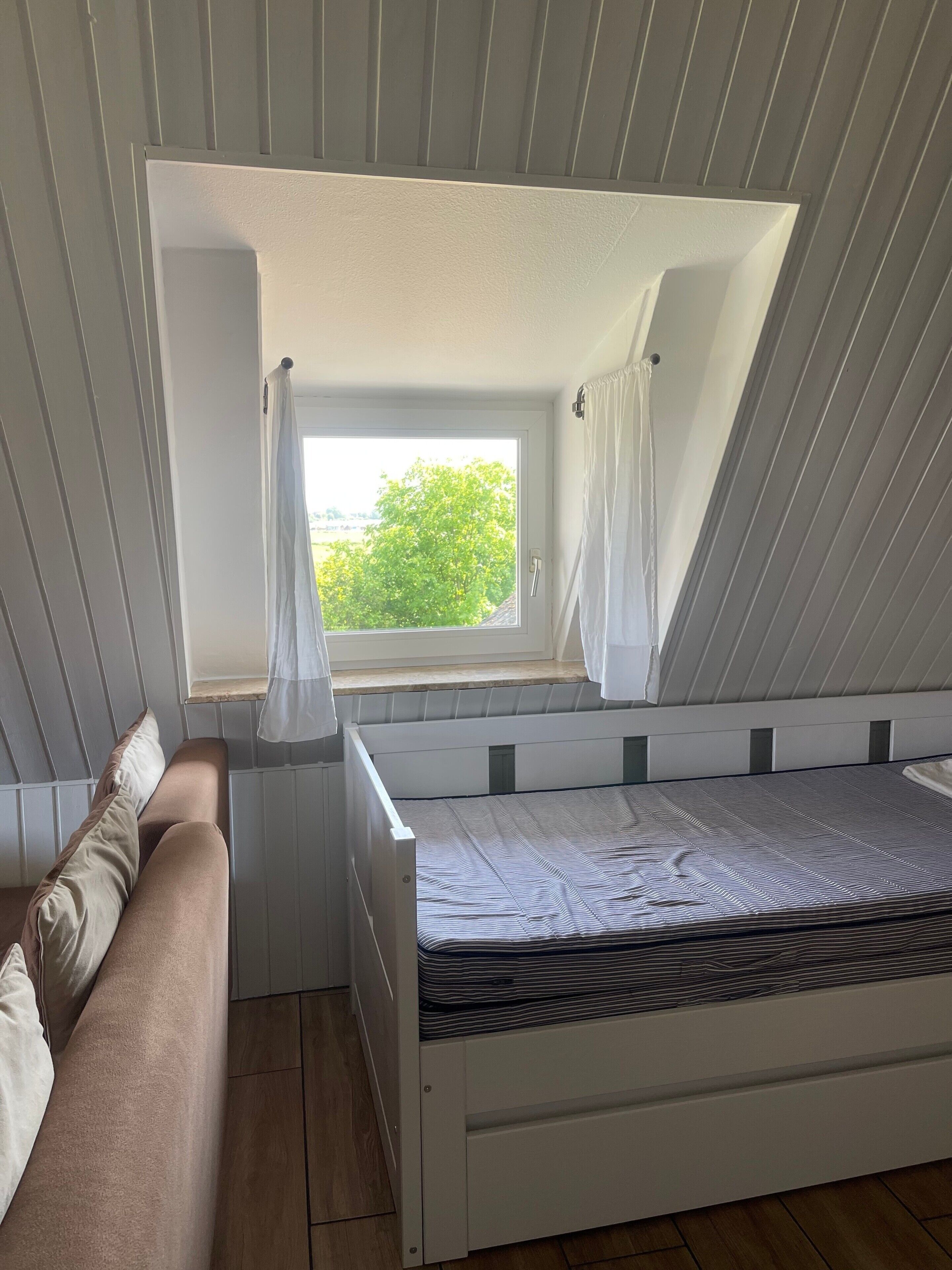 A small, single-bed room with a sloped ceiling, a window with curtains, and a wooden floor.