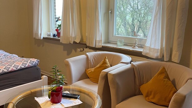 Una habitación con cama, dos sillones, una mesa redonda y un ventanal con cortinas.